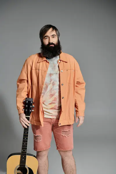 A man stands against a grey background, holding a guitar. He is dressed in a coral-colored jacket, shorts, and a tie-dye shirt. — Stock Photo