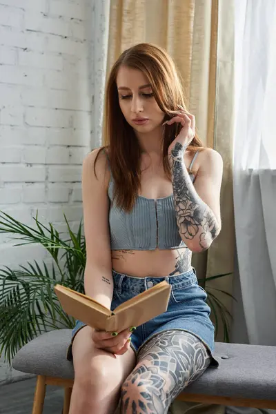 A young woman with tattoos sits on a bench, reading a book in her modern apartment. — Stock Photo