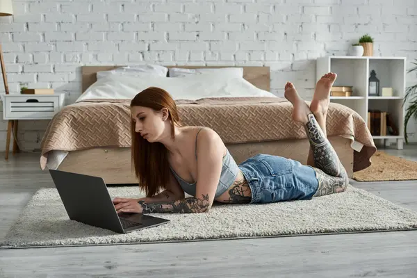 Eine junge tätowierte Frau liegt auf einem Plüschteppich in einer modernen Wohnung und arbeitet an ihrem Laptop. — Stockfoto