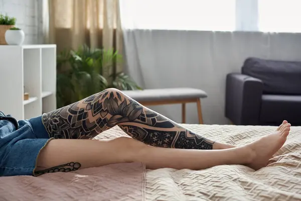 A woman with a leg tattoo relaxes on a bed in a modern apartment, enjoying a cozy weekend at home. — Stock Photo