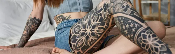 A young woman with intricate tattoos on her arm and leg relaxes on a bed in a modern apartment. — Stock Photo