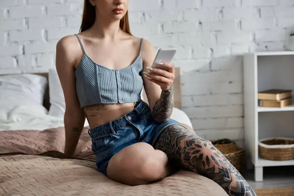 Uma jovem com tatuagens relaxa em uma cama em seu apartamento moderno, rolando casualmente através de seu telefone. — Stock Photo