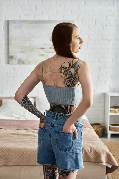 A young woman with tattoos on her back and arms stands in her modern apartment, hands in her pockets, looking out the window. — Stock Photo