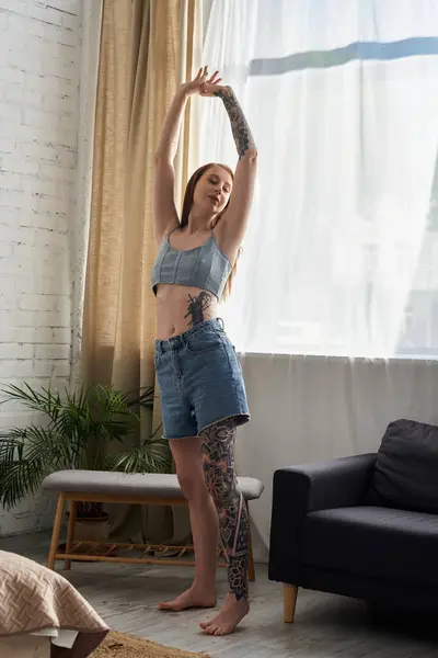 A young woman with tattoos stretches in a modern apartment, enjoying a relaxing weekend at home. — Stock Photo