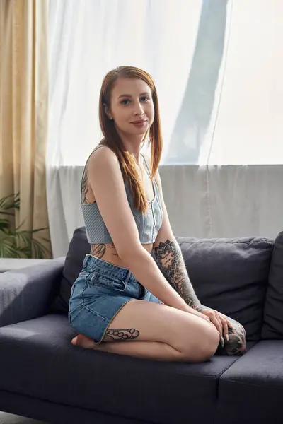 A young woman with tattoos sits on a couch in her modern apartment. She is enjoying a relaxing weekend at home. — Stock Photo