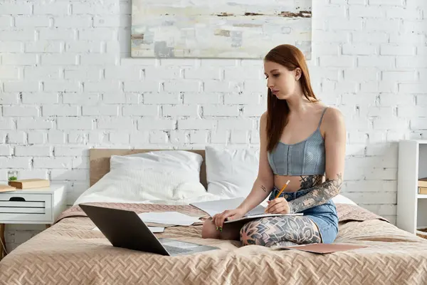 A young woman with tattoos sits on a bed in a modern apartment, sketching in a notebook on a cozy weekend. — Stock Photo