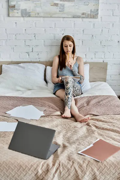 A young woman with tattoos sits on her bed in a modern apartment, thoughtfully writing in a notebook. — Stock Photo