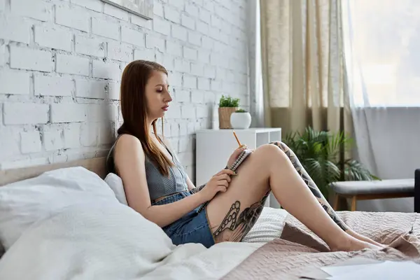 Eine junge Frau mit Tätowierungen sitzt auf ihrem Bett in einer modernen Wohnung und schreibt in ein Notizbuch. — Stockfoto