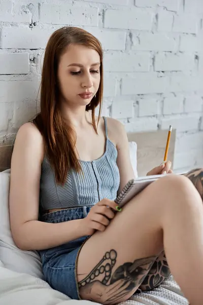 A young woman with tattoos sits on a bed in a modern apartment, writing in a notebook. — Stock Photo