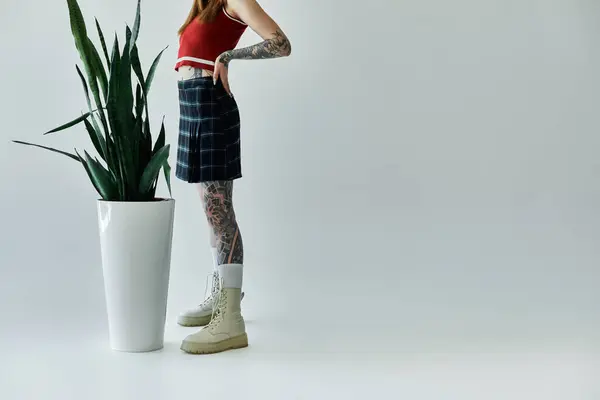 Una joven con tatuajes lleva una camiseta roja y una falda a cuadros, de pie frente a un fondo gris. - foto de stock