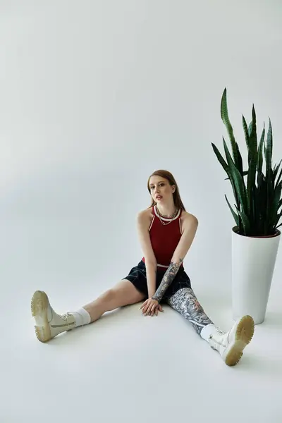 A young woman with tattoos sits on a grey background, wearing a red tank top and plaid skirt. — Stock Photo