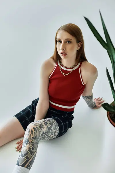 Una joven con tatuajes se sienta sobre un fondo blanco con una camiseta roja y una falda a cuadros. - foto de stock