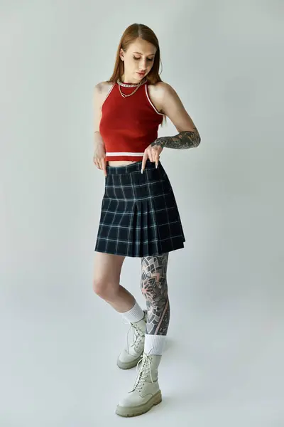 A young woman with tattoos stands in a plaid skirt and red tank top, her legs crossed at the ankles. — Stock Photo