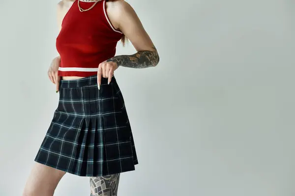 Uma jovem mulher com tatuagens em seus braços posa em um top de tanque vermelho e uma saia xadrez contra um fundo cinza. — Fotografia de Stock