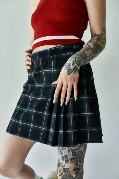 A tattooed young woman in a plaid skirt and tank top poses against a grey background. — Stock Photo