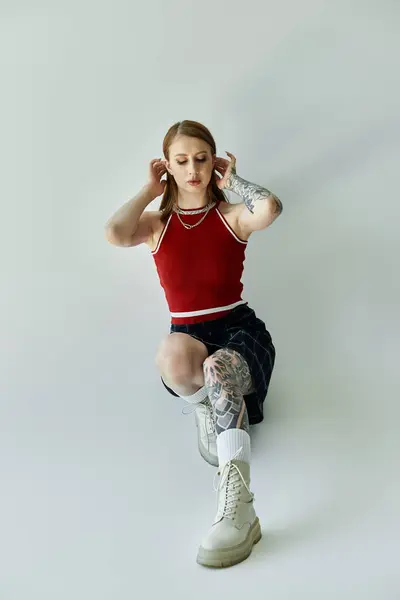 A young woman with tattoos sits on a grey background, wearing a red tank top, plaid skirt, and white boots. — Stock Photo