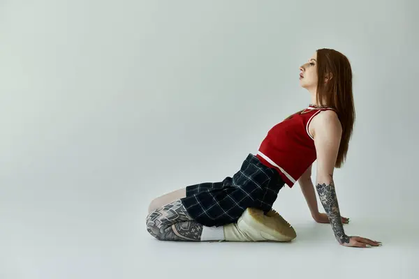 A tattooed young woman wearing a red tank top and a plaid skirt poses on a grey background. — Stock Photo