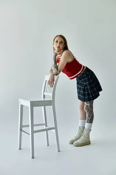 A young woman with tattoos stands leaning on a white chair, wearing a red tank top and a plaid skirt. — Stock Photo