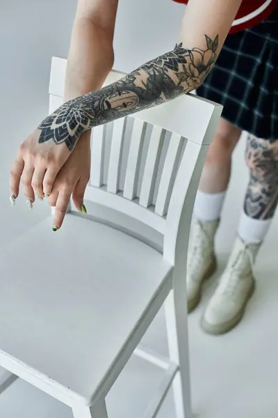 A young woman with tattoos on her arm leans on a white chair. She is wearing a plaid skirt — Stock Photo