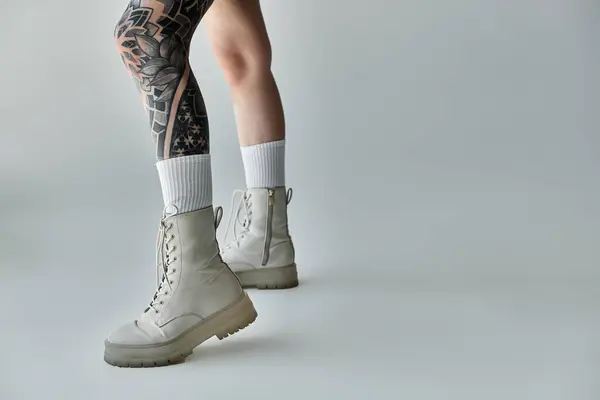 Un primo piano di una donna tatuata gambe, in mostra stivali bianchi e calzini bianchi su uno sfondo grigio. — Stock Photo