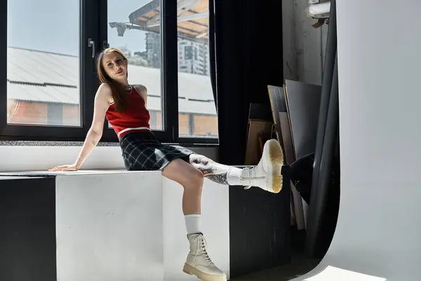 A young woman with tattoos sits on a window ledge wearing a red tank top and a plaid skirt. — Stock Photo