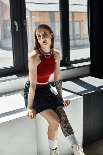 A young woman with tattoos poses in a red tank top and plaid skirt against a grey background. — Stock Photo