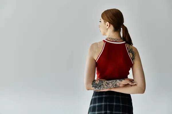 A young woman with tattoos on her arms wears a red tank top and plaid skirt, standing against a grey backdrop. — Stock Photo
