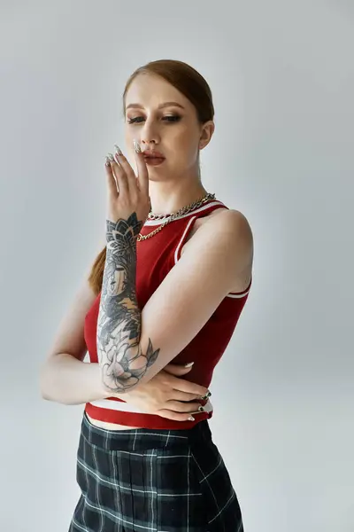 Una joven con tatuajes en el brazo posa sobre un fondo gris, llevando una falda a cuadros y una camiseta roja. - foto de stock