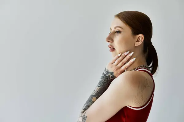 A young woman with tattoos on her arm poses in a red tank top against a grey background. — Stock Photo