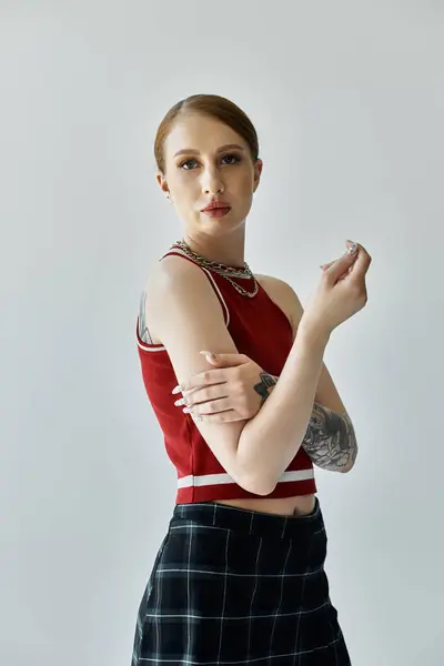 A young woman with tattoos poses in a red tank top and plaid skirt against a grey background. — Stock Photo