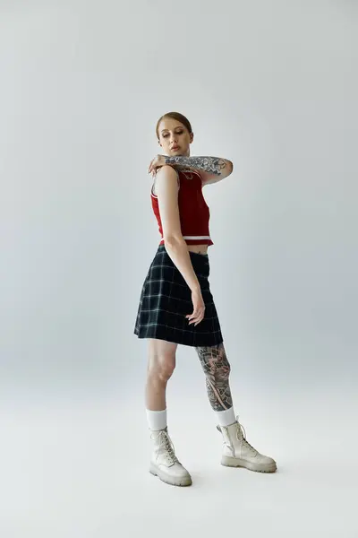 A young woman with tattoos stands in front of a grey backdrop, wearing a red tank top, black and white plaid skirt, and white boots. — Stock Photo