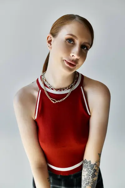 Una mujer joven con tatuajes con una camiseta roja y una falda a cuadros se levanta sobre un fondo gris. - foto de stock