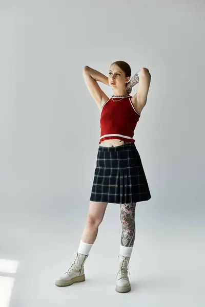 A young woman in a plaid skirt and red tank top poses against a grey background, showcasing her confident style. — Stock Photo