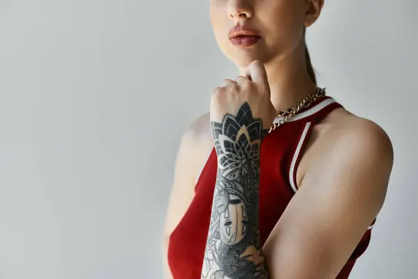 Una giovane donna con un braccio tatuato si erge su uno sfondo grigio, indossando una canotta rossa e una catena d'oro. — Foto stock