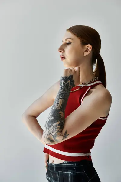 Una joven con un tatuaje de manga completa se levanta sobre un fondo gris, vistiendo una camiseta roja y falda a cuadros. - foto de stock