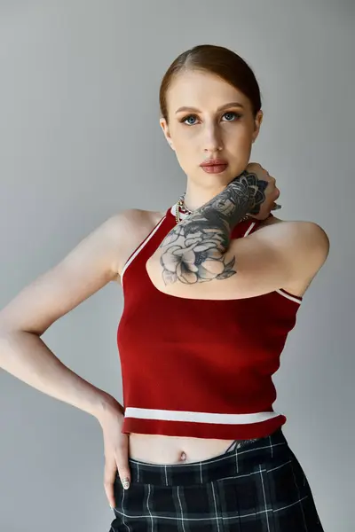 Eine junge Frau mit Tätowierungen am Arm, mit rotem Tanktop und kariertem Rock, posiert vor grauem Hintergrund. — Stockfoto
