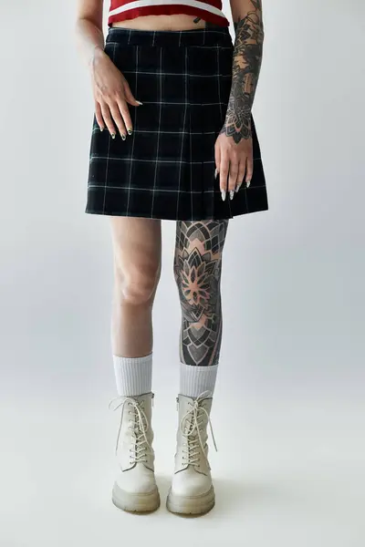 A young woman in a plaid skirt and white boots stands against a grey background. Her leg is adorned with a large tattoo. — Stock Photo