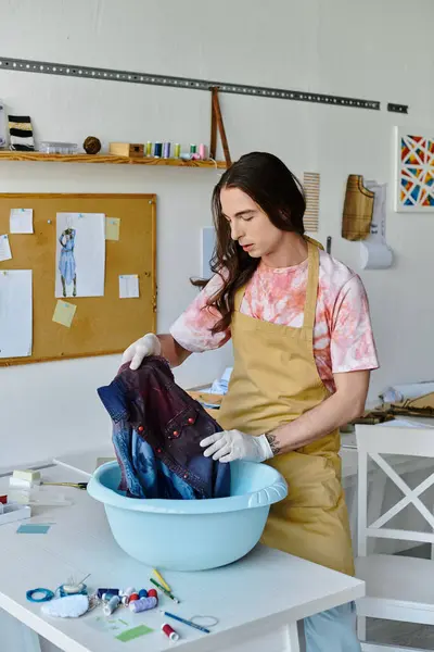 A young man wearing a tie-dye shirt and a yellow apron dyes a piece of clothing in his atelier. — Stock Photo