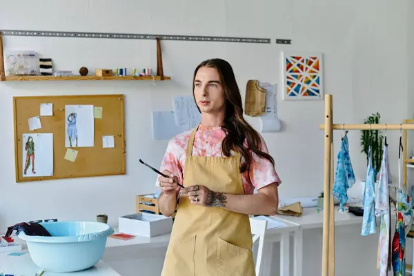 A young man stands in his clothing restoration atelier, holding a paintbrush, surrounded by his creative work. — Stock Photo