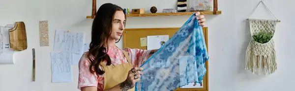 A young man in a light pink t-shirt and apron holds up a vibrant blue and white tie-dye fabric in his clothing restoration atelier. — Stock Photo