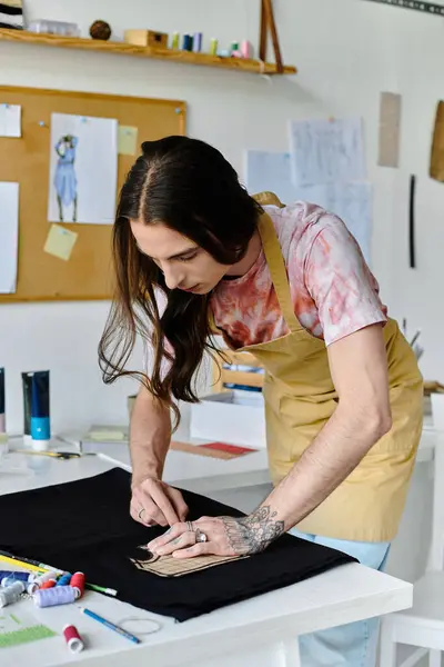 A young man with long brown hair is meticulously working on a black garment, surrounded by various sewing supplies. — Stock Photo