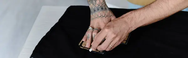 A young man with a tattoo on his forearm carefully repairs a piece of clothing in his DIY clothing restoration atelier. — Stock Photo