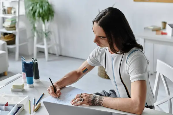 Um jovem com cabelos longos senta-se em uma mesa em seu atelier, esboçando desenhos em um pedaço de papel. — Fotografia de Stock