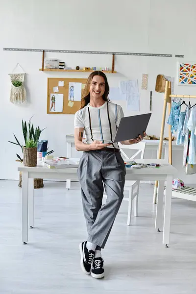 A young man stands in his clothing restoration atelier, laptop in hand, showcasing his passion for sustainable fashion and giving new life to discarded clothing. — Stock Photo