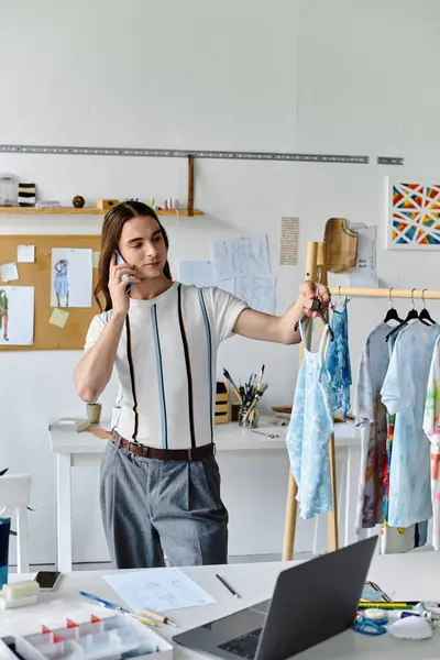 Un joven gay en un taller de restauración de ropa de bricolaje, por teléfono, mostrando una prenda que ha sido reutilizada para una línea de ropa sostenible. - foto de stock