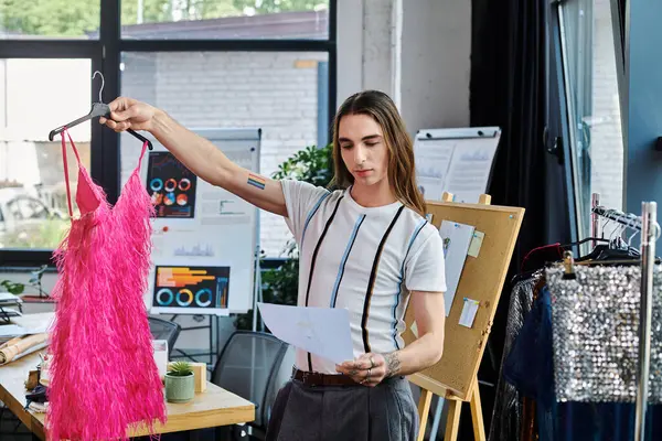 Молодой гей осматривает розовое пернатое платье в мастерской по реставрации одежды, подчеркивая свою приверженность к устойчивой моде и давая новую жизнь старым предметам. — стоковое фото