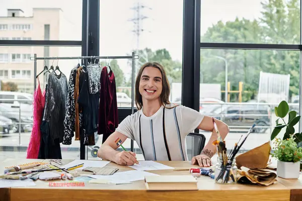 A young gay man smiles as he sketches designs in his DIY clothing restoration atelier. He is surrounded by colorful fabrics and tools, showcasing his passion for sustainable fashion. — Stock Photo