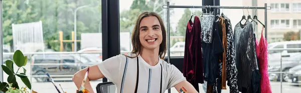 Um jovem gay sorri confiantemente em seu atelier de restauração de roupas, cercado por roupas aguardando transformação. — Fotografia de Stock