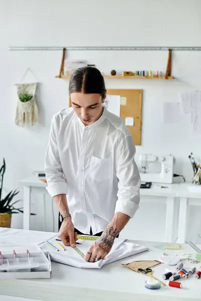 A young man in a white shirt measures fabric with a tape measure, meticulously working on a new clothing project in his sustainable atelier. — Stock Photo