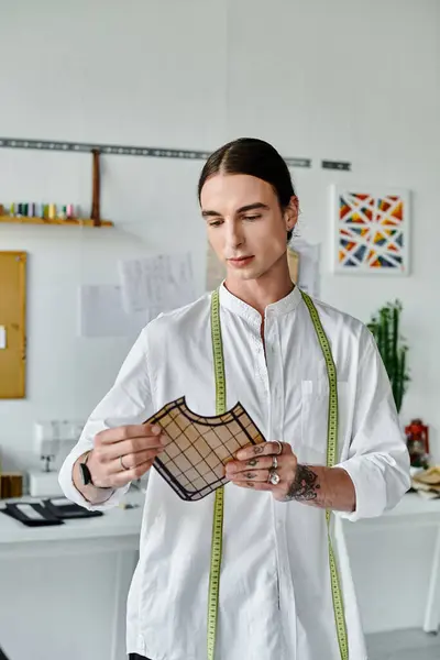 A young man in a white shirt, with a measuring tape around his neck, examines a piece of fabric in his clothing restoration atelier. — Stock Photo